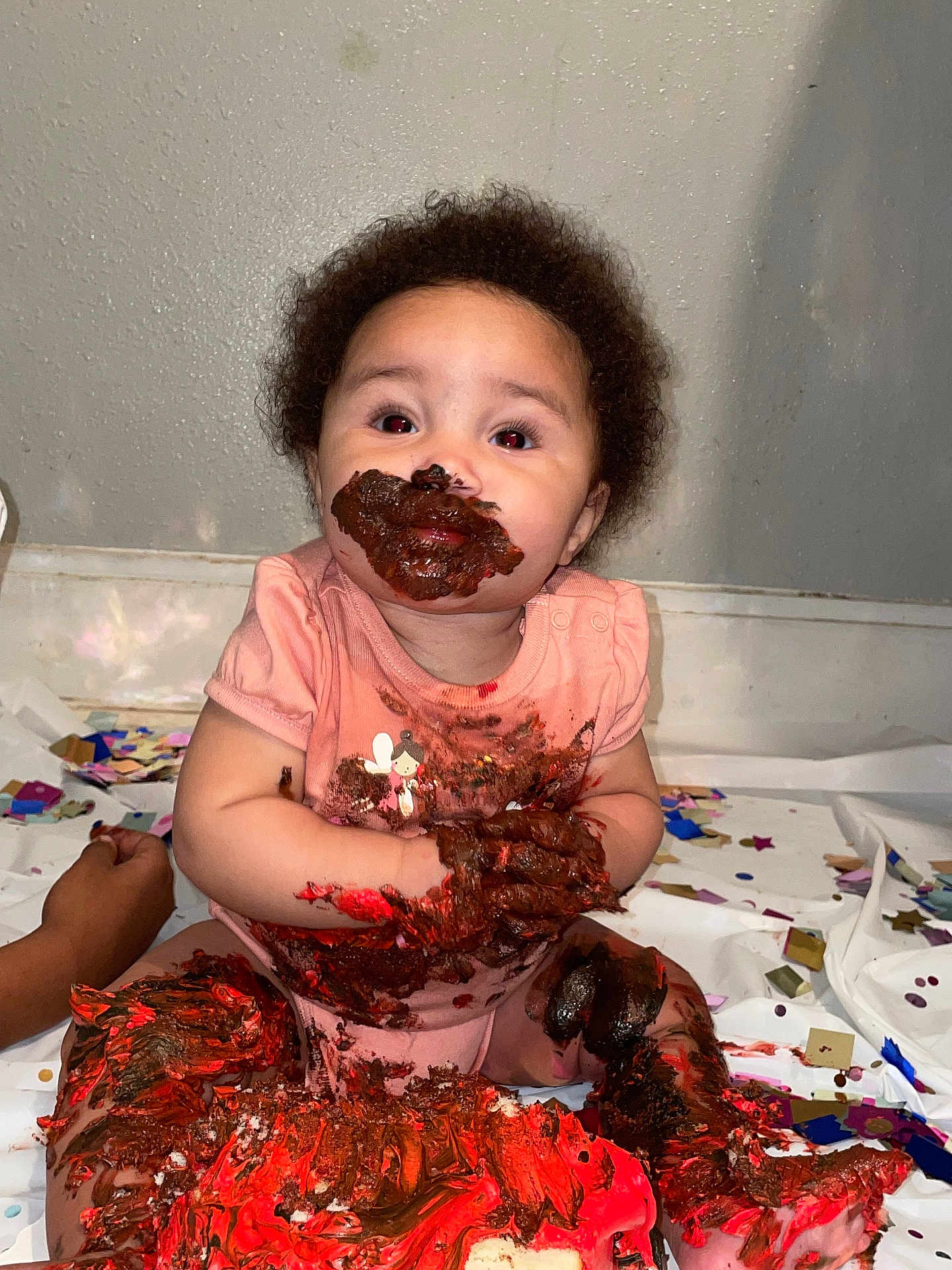 Kihanna is registered to the contest to win money with this photo: baby, cake, chocolate, messy, confetti, celebration, face, hands, legs, pink_clothing, sitting, smile, party, indoor, wall, floor, food, fun, child, happy