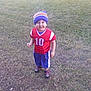 child, toddler, smiling, grass, outdoor, beanie, hat, red_jersey, blue_shorts, sporty_clothing, shoes, number_10, happy, young_child, standing, casual, daylight, nature, grass_field, portrait