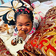 Maddison is registered to the contest to win money with this photo: baby, child, smiling, curly_hair, red_bows, toy, hand, cushion, pillow, patterned_fabric, butterfly, bed, blanket, indoor, playful, happy, face, person, closeup, cozy