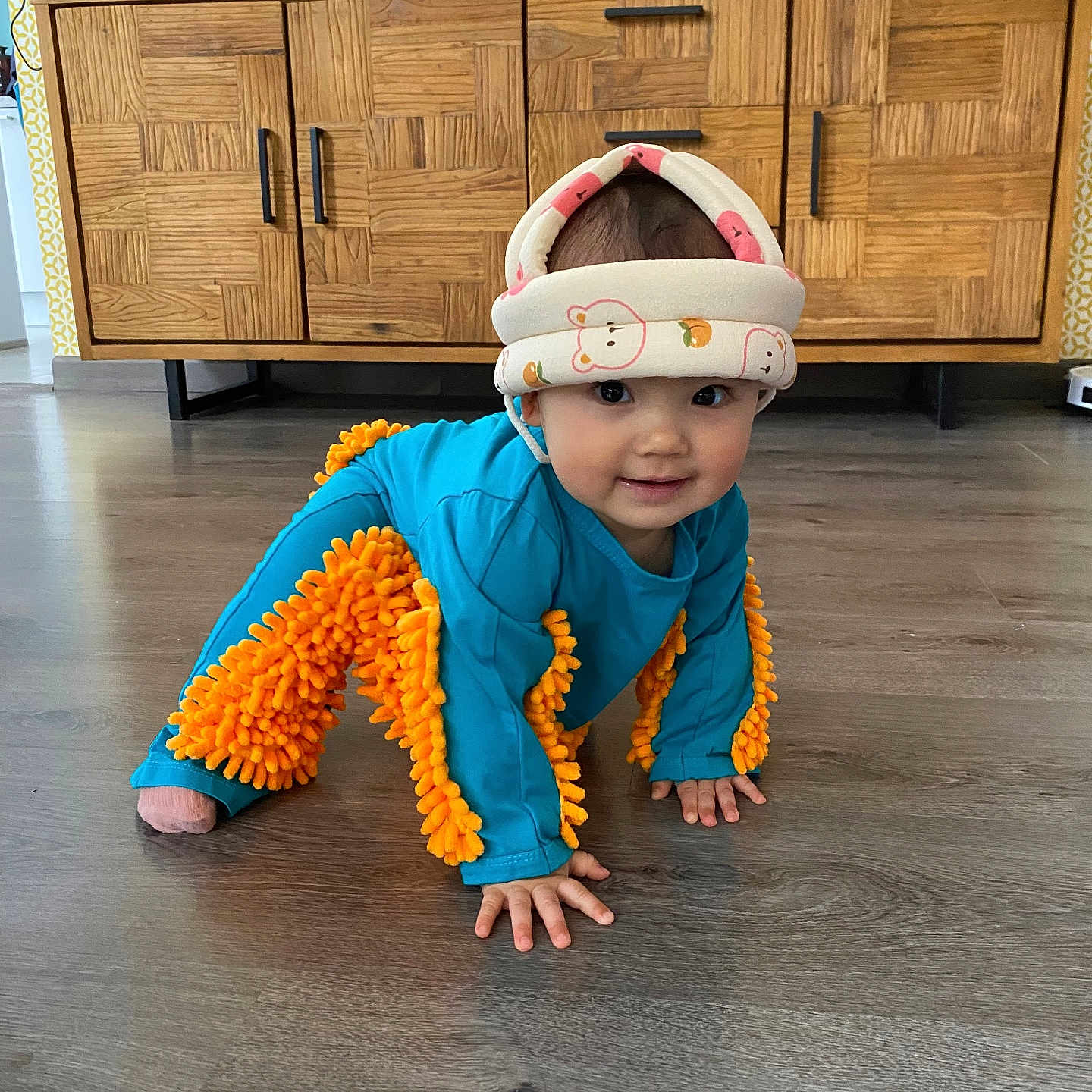 Lucie Minh Lam a rejoint le concours — aidez-le/la à gagner de superbes lots ! baby, blue_outfit, cabinet, child, crawling, cute, floor, furniture, headwear, helmet, home, household_items, indoor, orange_fluffy_accent, person, playful, smiling, toddler, wooden_floor, young_child