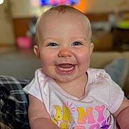Presley is registered to the contest to win money with this photo: baby, smiling, blue_eyes, indoor, shirt, colorful_text, happy, child, portrait, cute, infant, skin, face, sitting, person, human, young, cheerful, closeup, warm_lighting