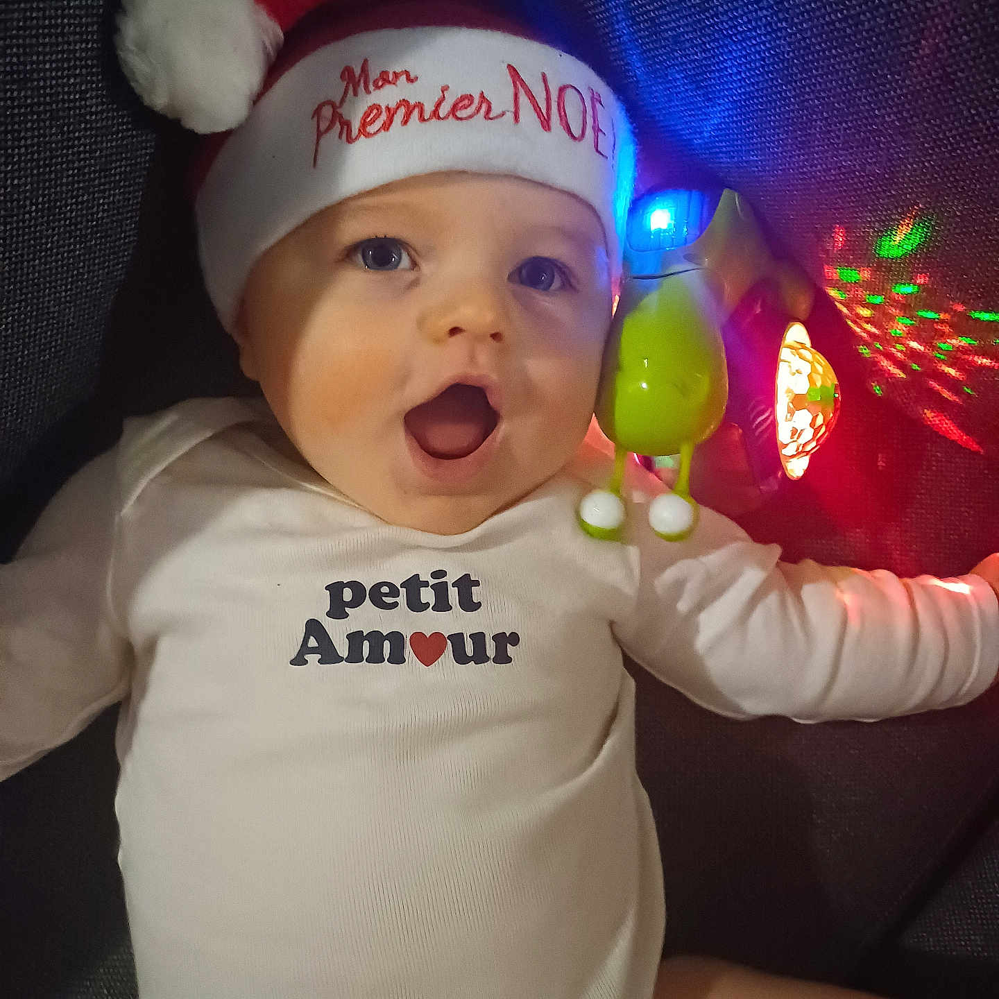 Aaron a rejoint le concours — aidez-le/la à gagner de superbes lots ! baby, child, christmas, closeup, colorful_lights, cute, expression, face, festive, glowing, happy, holiday, indoor, infant, lying_down, onesie, portrait, santa_hat, text_clothing, toy