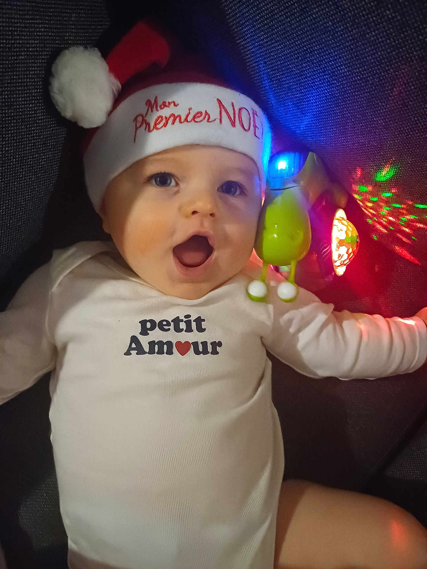 Aaron a rejoint le concours — aidez-le/la à gagner de superbes lots ! baby, infant, child, santa_hat, christmas, holiday, onesie, text_clothing, toy, glowing, colorful_lights, face, expression, happy, cute, lying_down, indoor, closeup, portrait, festive