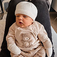 Ezra a rejoint le concours — aidez-le/la à gagner de superbes lots ! baby, infant, knitted_hat, mittens, beige_clothing, teddy_bear_design, person, lap, indoor, comfortable, resting, soft_fabric, pants, seated, cute, child, newborn, warm_lighting, cozy, casual