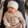 baby, infant, knitted_hat, mittens, beige_clothing, teddy_bear_design, person, lap, indoor, comfortable, resting, soft_fabric, pants, seated, cute, child, newborn, warm_lighting, cozy, casual