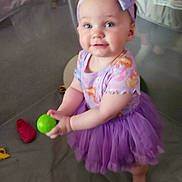Raven is registered to the contest to win money with this photo: baby, barefoot, child, childhood, clothing, cute, floor, happy, headband, holding_object, indoor, infant, person, play_area, playpen, purple_tutu, smiling, toddler, toy_ball, young_child