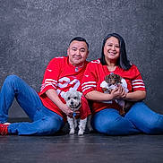 Hermione is registered to the contest to win money with this photo: man, woman, dog, pet, jeans, red_clothing, sports_jersey, sitting, smiling, indoor, studio, floor, casual, short_hair, portrait, couple, paw, small_dog, dachshund, white_dog