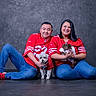 man, woman, dog, pet, jeans, red_clothing, sports_jersey, sitting, smiling, indoor, studio, floor, casual, short_hair, portrait, couple, paw, small_dog, dachshund, white_dog