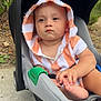 baby, child, car_seat, hooded_towel, orange_stripes, outdoor, sidewalk, greenery, hand, foot, curious, toddler, person, infant, seat, cute, casual, daylight, portrait, expression