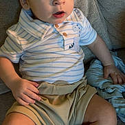 Remington is registered to the contest to win money with this photo: baby, child, infant, sitting, striped_shirt, dinosaur_patch, shorts, couch, furniture, indoors, curious, face, hand, legs, casual_clothing, young_child, comfort, expression, human, portrait
