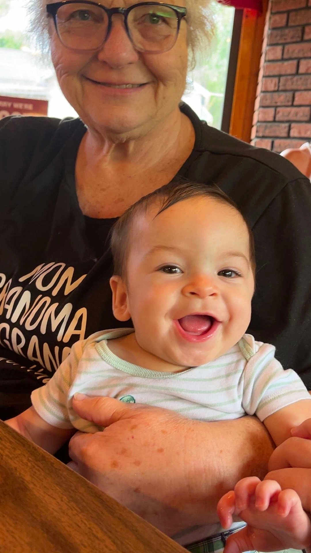 Remington joined the competition — help win amazing prizes! baby, smiling, woman, glasses, indoor, holding, table, brick_wall, shirt, text_on_shirt, happy, person, face, hand, seated, light, skin, portrait, family, cozy