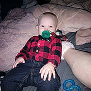 Justin is registered to the contest to win money with this photo: baby, infant, child, portrait, sitting, blanket, bed, pajamas, checkered_shirt, red_clothing, black_clothing, jeans, pacifier, toy, blue_toy, bedroom, indoors, face, wide_eyes, cozy