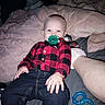 baby, infant, child, portrait, sitting, blanket, bed, pajamas, checkered_shirt, red_clothing, black_clothing, jeans, pacifier, toy, blue_toy, bedroom, indoors, face, wide_eyes, cozy