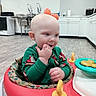 Justin is registered to the contest to win money with this photo: baby, infant, child, portrait, sitting, smiling, curious, hand_in_mouth, green_outfit, festive_clothing, orange_toy, activity_center, baby_walker, playroom, kitchen, home, indoors, colorful_toys, wood_floor, close_up