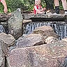 baby, child, waterfall, rocks, outdoors, nature, stream, forest, red_outfit, sitting, adult, legs, shoes, socks, foliage, stone, water, park, daylight, landscape