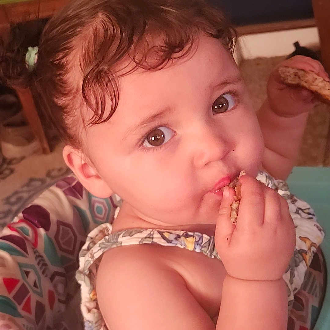 Tiya is registered to the contest to win money with this photo: baby, bread, brown_eyes, child, close_up, colorful, curly_hair, cute, eating, expression, face, hand, indoor, patterned, person, portrait, seat, skin, snack, toddler