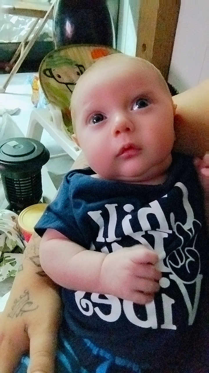 Shane is registered to the contest to win money with this photo: baby, infant, person, face, arm, tattoo, clothing, indoor, chair, expression, looking_up, skin, hand, seated, casual, young_child, curious, blue_shirt, closeup, human