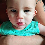 Apollo joined the competition — help win amazing prizes! baby, infant, child, portrait, close_up, face, wide_eyes, looking_at_camera, turquoise_shirt, clothing, adult_arm, tattoo, indoor, seated, supported, head, eyes, mouth, skin, photography
