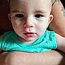 baby, infant, child, portrait, close_up, face, wide_eyes, looking_at_camera, turquoise_shirt, clothing, adult_arm, tattoo, indoor, seated, supported, head, eyes, mouth, skin, photography