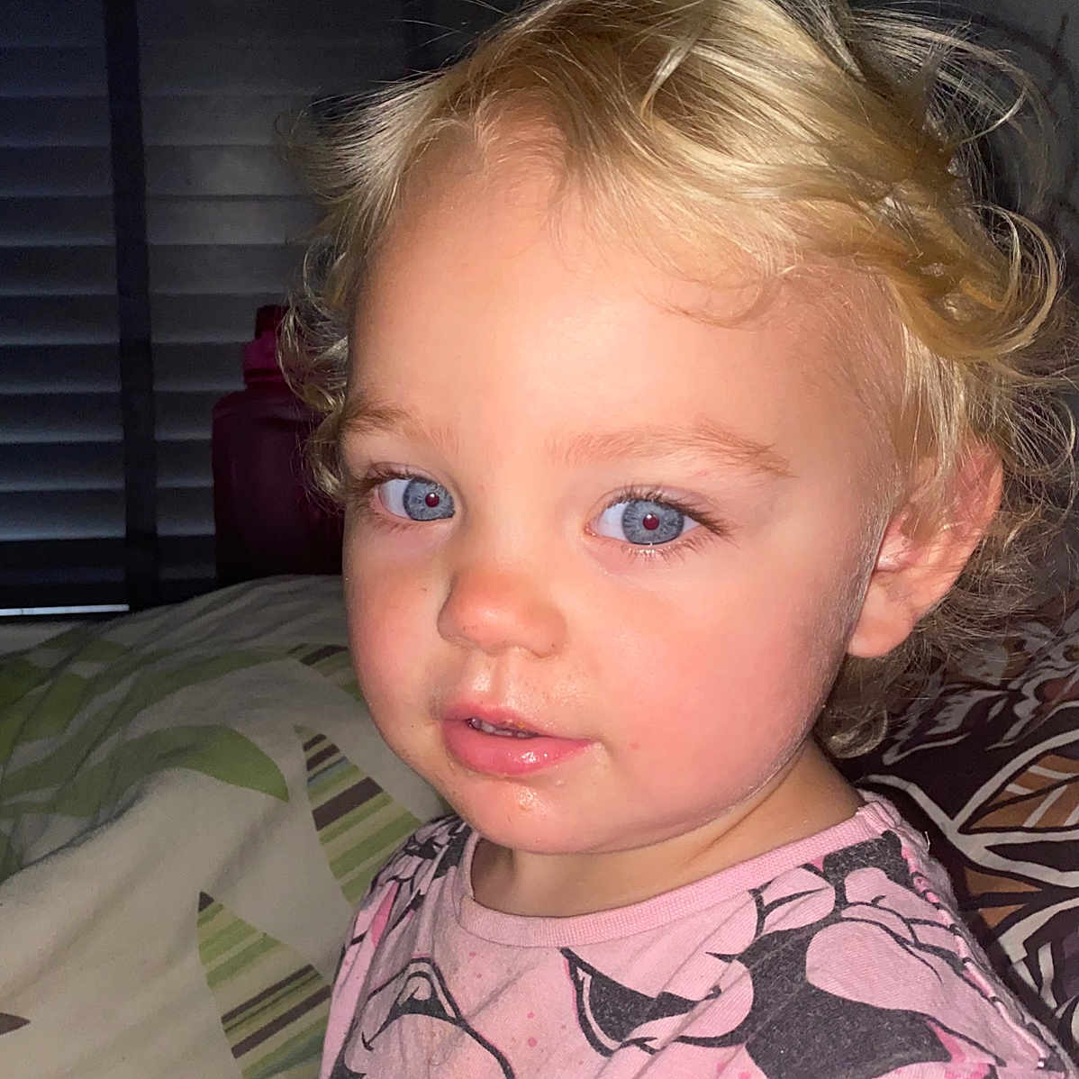 Kyra joined the competition — help win amazing prizes! bed, blanket, blonde_hair, blue_eyes, cartoon_print, casual_clothing, child, closeup, curious_expression, curly_hair, face, flash_photography, indoor, messy_face, person, pink_shirt, portrait, sitting, toddler, young_child