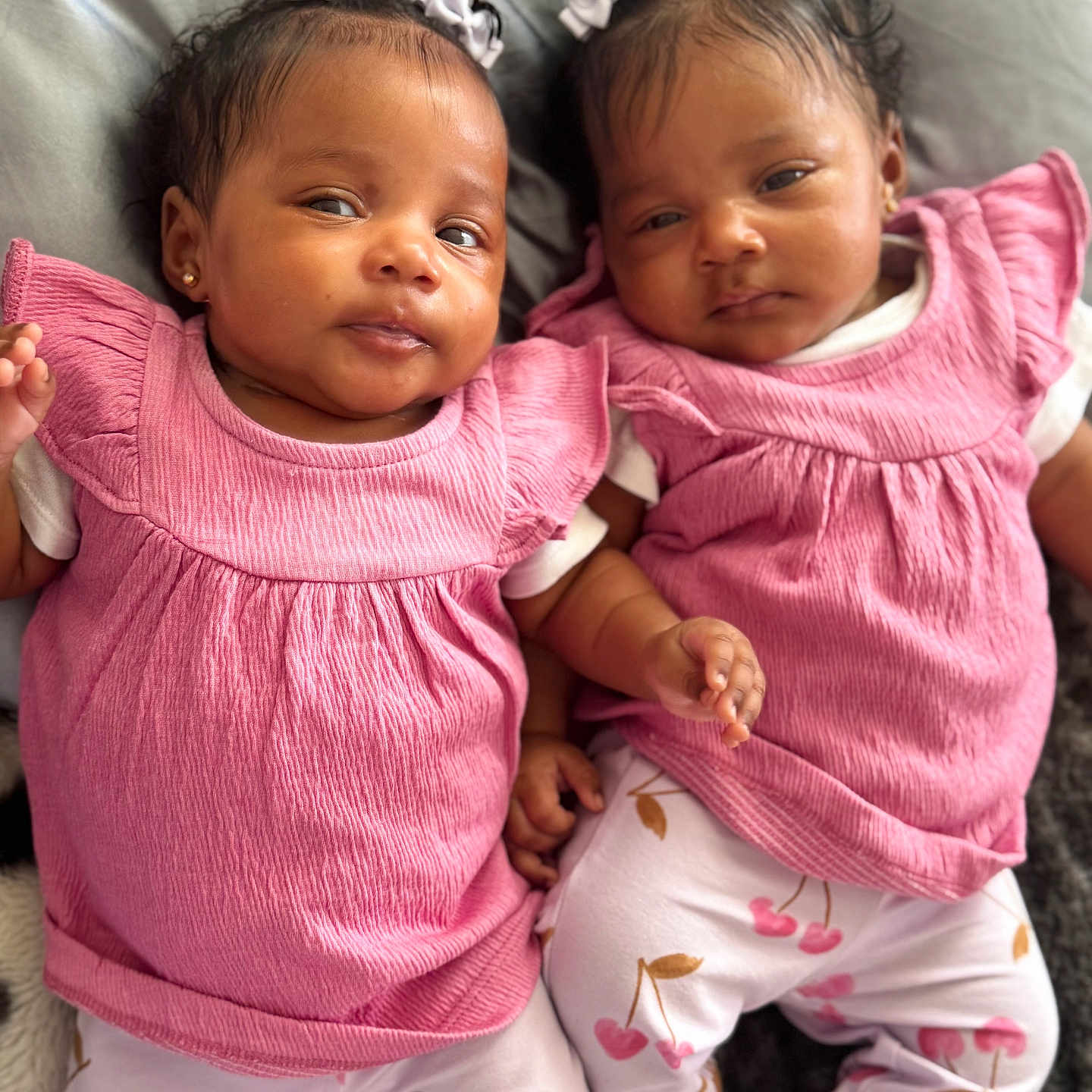 Trinity And Serenity is registered to the contest to win money with this photo: adorable, baby, cherry_pattern, child, cute, face, hair_bow, head, indoor, infant, lying_down, matching_outfits, person, pink_clothing, portrait, skin, small_hand, soft_surface, twin, white_socks