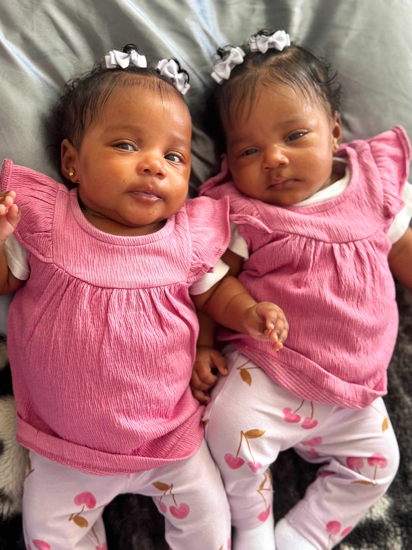 Trinity And Serenity is registered to the contest to win money with this photo: baby, twin, infant, pink_clothing, hair_bow, lying_down, cute, child, face, person, matching_outfits, white_socks, cherry_pattern, soft_surface, adorable, portrait, indoor, skin, head, small_hand