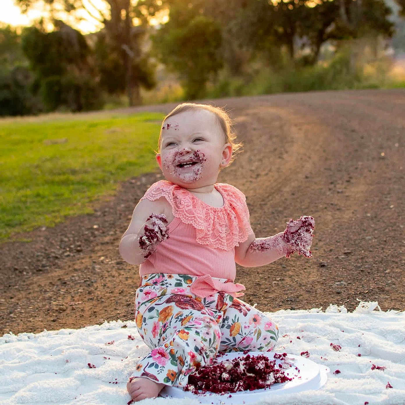 Wayah joined the competition — help win amazing prizes! baby, blanket, cake, celebration, child, dirt_path, face, floral_pants, food, grass, hands, happy, messy, nature, outdoor, pink_top, portrait, sitting, smiling, sunset
