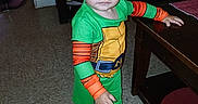 Benjamin is registered to the contest to win money with this photo: cabinet, child, costume, curious, curly_hair, floor, green_clothing, home, indoor, living_room, orange_clothing, person, socks, standing, table, television, toddler, wide_eyes, wooden_furniture, young_child
