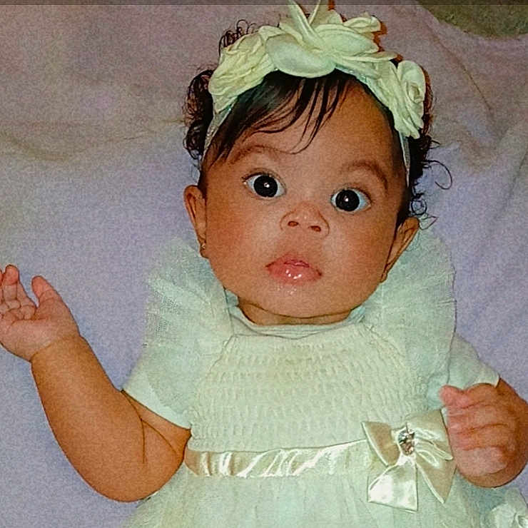 Emarelys is registered to the contest to win money with this photo: baby, child, dress, headband, flower, wide_eyes, curious, portrait, indoor, cute, infant, person, skin, hand, sleeve, ribbon, fabric, young, face, expression