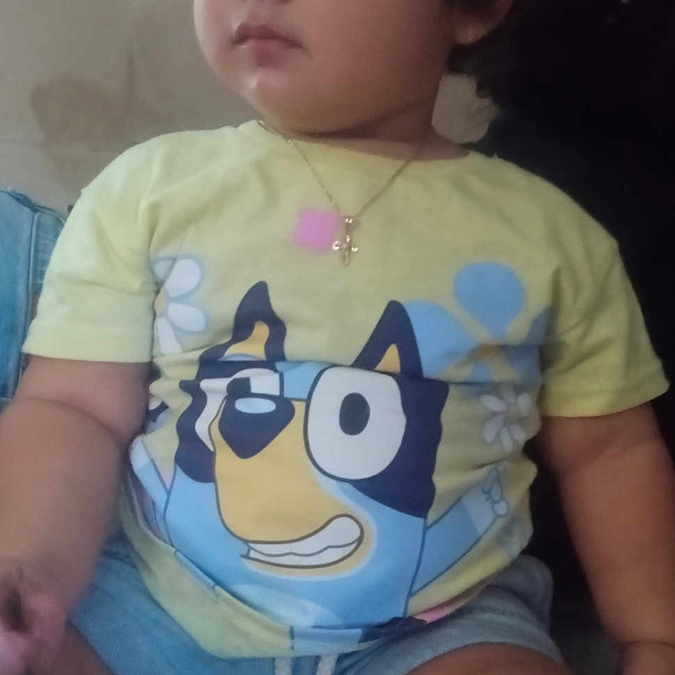 Juno is registered to the contest to win money with this photo: baby, cartoon_print, casual_clothing, child, curly_hair, cute, denim_shorts, expression, footwear, hand, indoors, leg, necklace, person, portrait, sitting, skin, soft_surface, toddler, yellow_tshirt