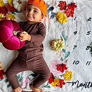 Avi participe au concours pour gagner de l'argent avec cette photo : baby, child, orange_hat, brown_clothing, plush_pumpkin, white_blanket, autumn_leaves, decor, numbers, months_marker, smile, face, hand, foot, lying_down, indoors, seasonal, cute, portrait, toddler