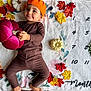 baby, child, orange_hat, brown_clothing, plush_pumpkin, white_blanket, autumn_leaves, decor, numbers, months_marker, smile, face, hand, foot, lying_down, indoors, seasonal, cute, portrait, toddler
