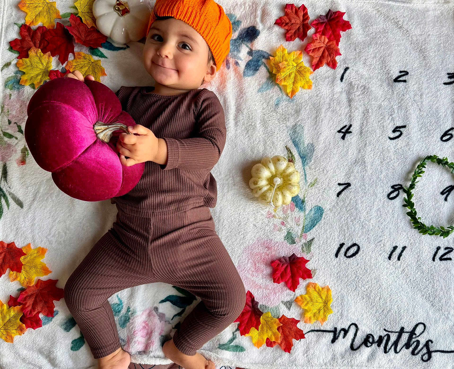 Avi participe au concours pour gagner de l'argent avec cette photo : baby, child, orange_hat, brown_clothing, plush_pumpkin, white_blanket, autumn_leaves, decor, numbers, months_marker, smile, face, hand, foot, lying_down, indoors, seasonal, cute, portrait, toddler