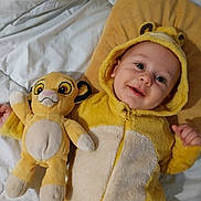 Mahé a rejoint le concours — aidez-le/la à gagner de superbes lots ! baby, child, plush_toy, yellow_onesie, bed, pillow, smile, happy, cute, infant, soft_toy, costume, hood, lying_down, indoor, person, toddler, animal_onesie, childrens_clothing, stuffed_animal