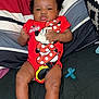baby, infant, onesie, minnie_mouse, red_clothing, socks, pacifier, pillow, striped_pillow, black_hair, curly_hair, indoor, couch, child, person, cute, smiling, relaxed, lying_down, portrait