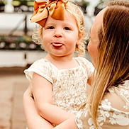 Diem joined the competition — help win amazing prizes! child, toddler, adult, holding, bow, orange_bow, playful, tongue_out, blond_hair, curly_hair, floral_clothing, person, outdoor, plants, potted_plants, blurred_background, portrait, cute, smile, affection