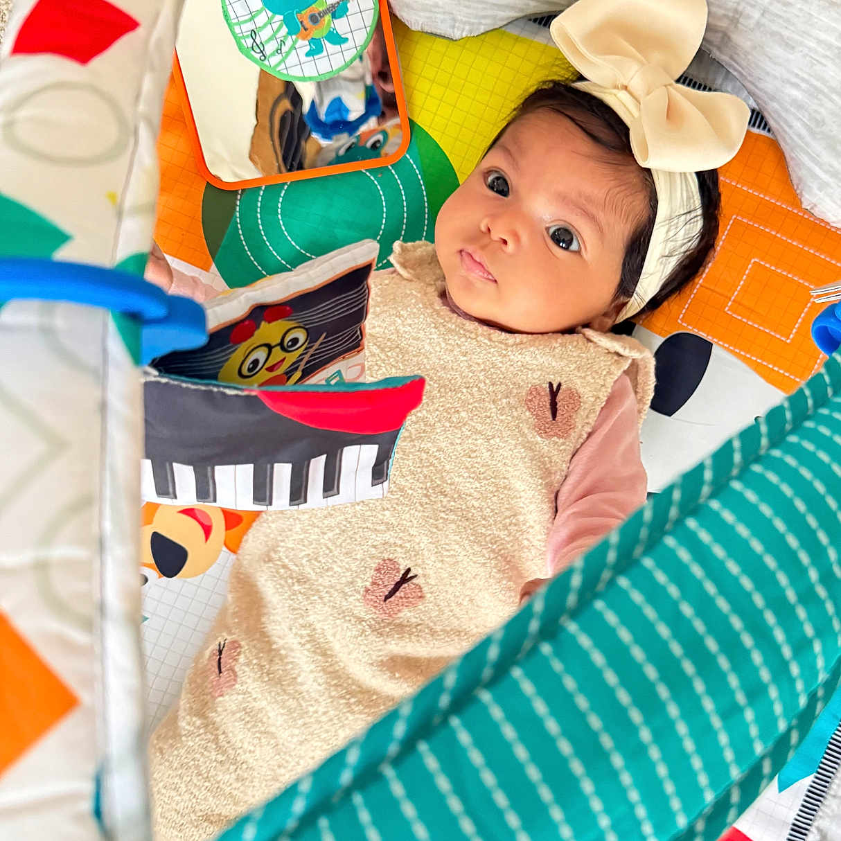 Joudia a rejoint le concours — aidez-le/la à gagner de superbes lots ! baby, bow, carpet, child, colorful, cute, expression, eyes, face, floor, headband, indoor, infant, looking_up, mirror, patterned_clothing, play_mat, soft_toys, toy, young