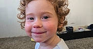 Sabrina joined the competition — help win amazing prizes! child, toddler, curly_hair, smile, indoor, carpet, face, person, white_shirt, rosy_cheeks, young_child, happy, cute, portrait, sitting, young_person, expression, head_and_shoulders, casual_clothing, home