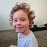 child, toddler, curly_hair, smile, indoor, carpet, face, person, white_shirt, rosy_cheeks, young_child, happy, cute, portrait, sitting, young_person, expression, head_and_shoulders, casual_clothing, home