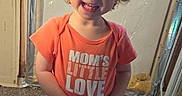 Sabrina is registered to the contest to win money with this photo: toddler, child, smiling, curly_hair, orange_shirt, text_on_clothing, barefoot, carpet, indoor, standing, playful, casual_clothing, young_child, happy, person, dinosaur_pants, home, wall, curly_hair_child, cute