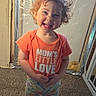 Sabrina is registered to the contest to win money with this photo: toddler, child, smiling, curly_hair, orange_shirt, text_on_clothing, barefoot, carpet, indoor, standing, playful, casual_clothing, young_child, happy, person, dinosaur_pants, home, wall, curly_hair_child, cute