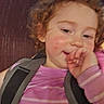 child, toddler, curly_hair, pink_shirt, striped_clothing, harness, smiling, hand_near_mouth, face, indoor, seat, casual, young_child, cute, portrait, person, expression, skin, hair, blurry
