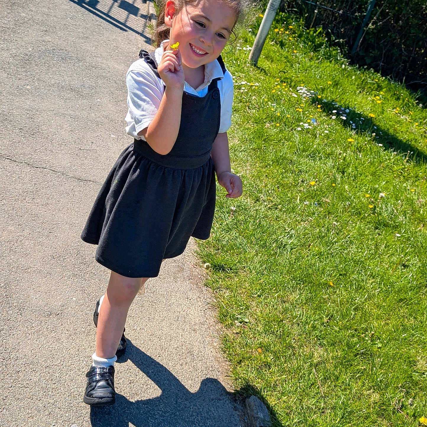 Melina is registered to the contest to win money with this photo: black_shoes, casual_clothing, child, daytime, dress, fence, flower, girl, grass, greenery, happy, nature, outdoor, pavement, person, portrait, shadow, smile, sunlight, white_socks