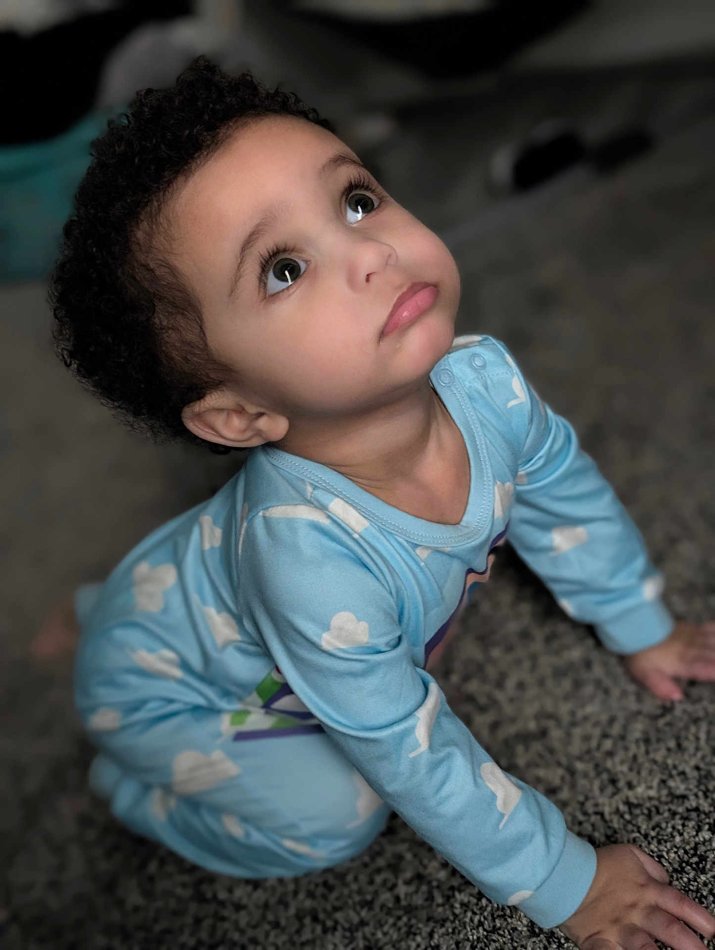 Everett is registered to the contest to win money with this photo: toddler, child, pajamas, blue_clothing, curly_hair, indoor, carpet, hands, face, expression, looking_up, cute, baby, person, young_child, eyes, skin, floor, clothing, long_sleeve