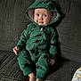 baby, child, green_onesie, dinosaur_pattern, hood, couch, sitting, barefoot, smiling, cute, infant, indoor, cozy, fabric, clothing, person, portrait, young_child, home, relaxed