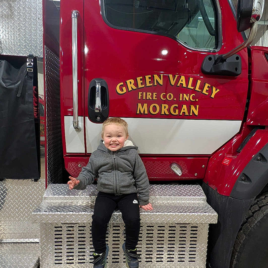 Cayden is registered to the contest to win money with this photo: boy, bumper, child, clothing, face, firetruck, footwear, head, machine, male, pants, person, photography, portrait, shoe, sitting, transportation, truck, vehicle, wheel
