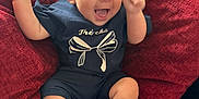 Annie is registered to the contest to win money with this photo: baby, child, smiling, happy, sitting, cushion, red_cushion, curly_hair, white_socks, navy_outfit, indoor, cute, joyful, person, infant, cheerful, raised_arms, comfortable, plush, portrait