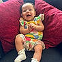 baby, smiling, child, colorful_clothing, white_socks, red_pillows, couch, indoor, happy, cute, infant, sitting, portrait, young_child, playful, cozy, fabric, home, person, expression
