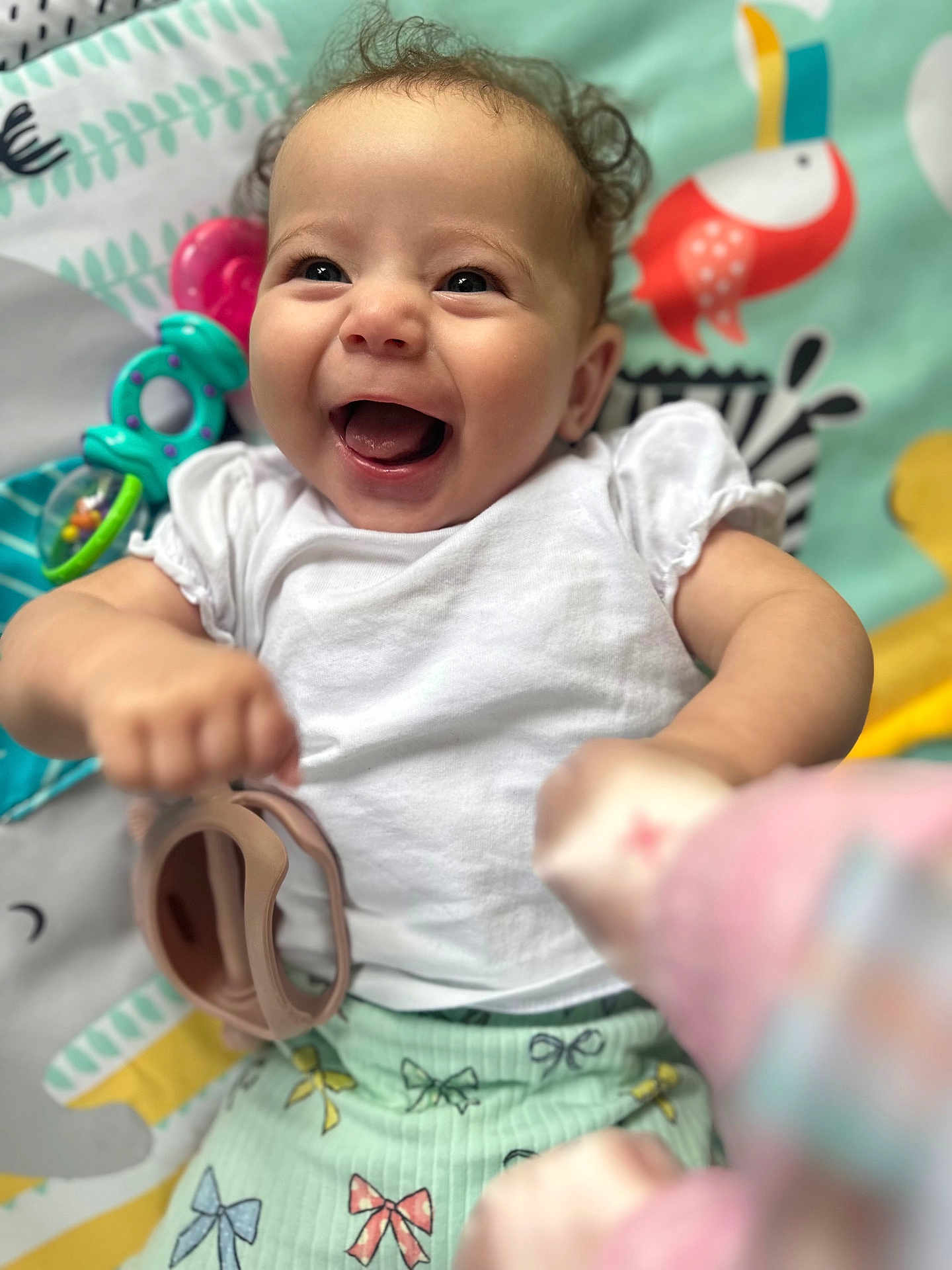 Annie joined the competition — help win amazing prizes! baby, smiling, child, white_shirt, play_mat, toy, happy, infant, curly_hair, close_up, colorful, patterned_clothing, cute, playful, cheerful, portrait, indoors, soft_lighting, hand, face