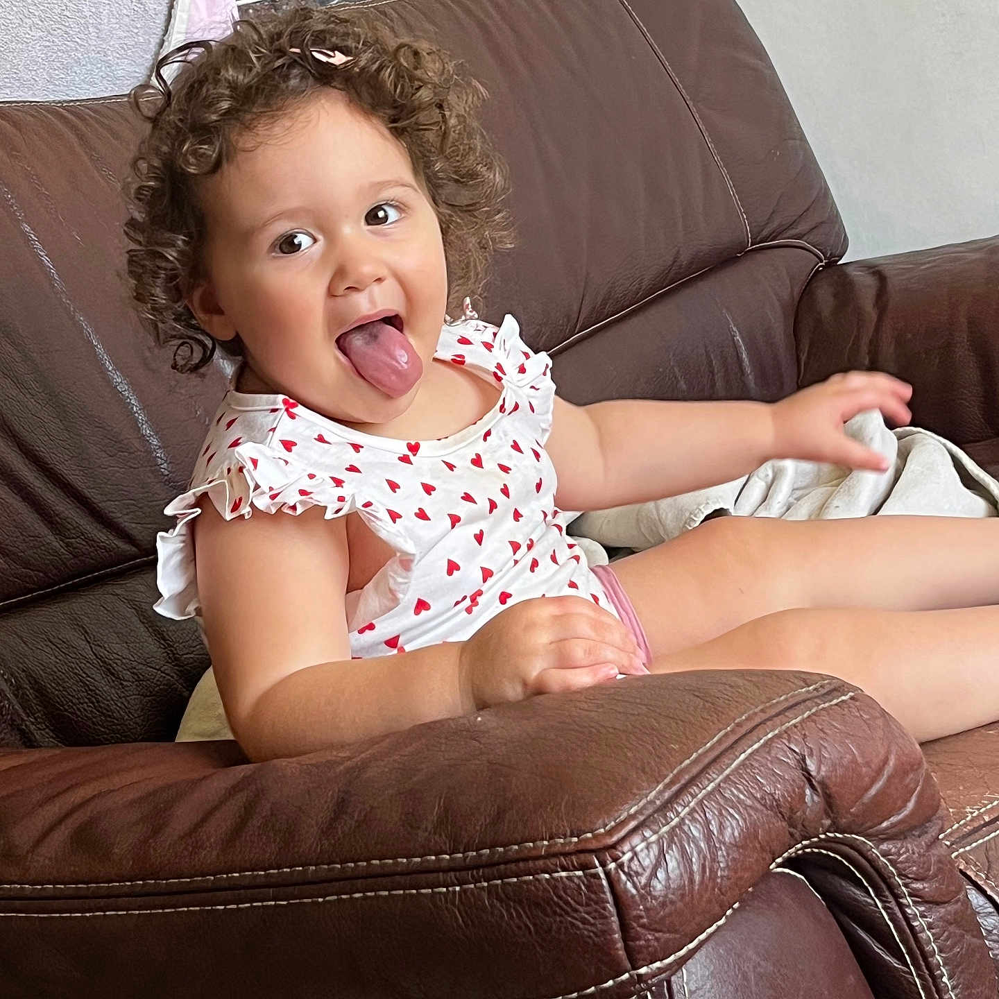 Juliette participe au concours pour gagner de l'argent avec cette photo : brown_couch, child, clothing, curly_hair, cute, expression, face, furniture, home, indoor, leather, person, playful, red_hearts, relaxed, seated, smiling, toddler, tongue_out, white_dress